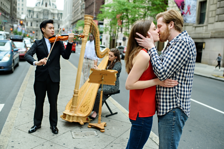 An engaged couple kiss on broad street with a harp and violin playing in the background.